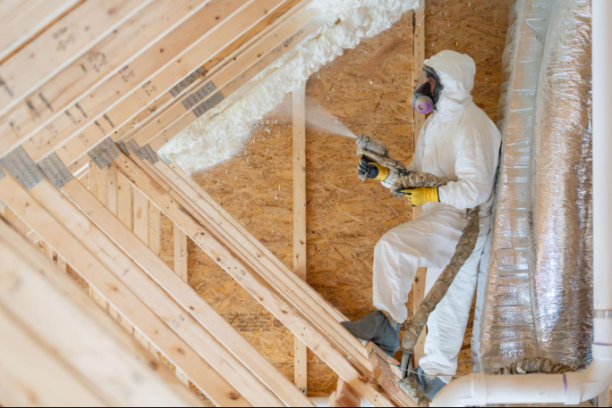 Worker in white protective suit insulating attic wall with a spray foam gun