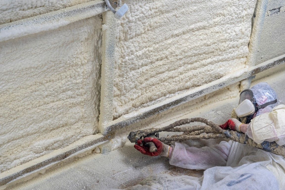 A worker in protective gear applies spray foam insulation to a wall using a spray gun.
