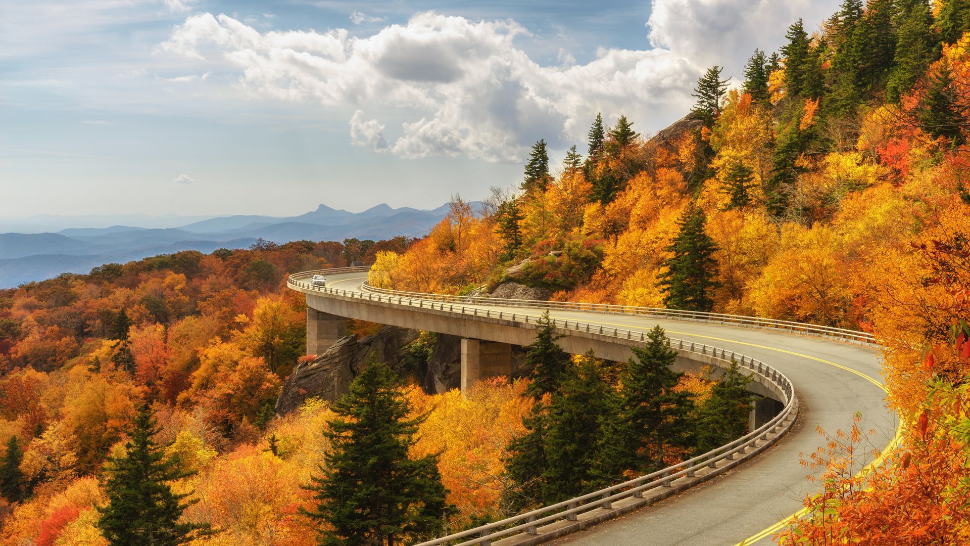 A curved bridge winds through a mountain forest with vibrant autumn foliage in shades of orange, yellow, and green.