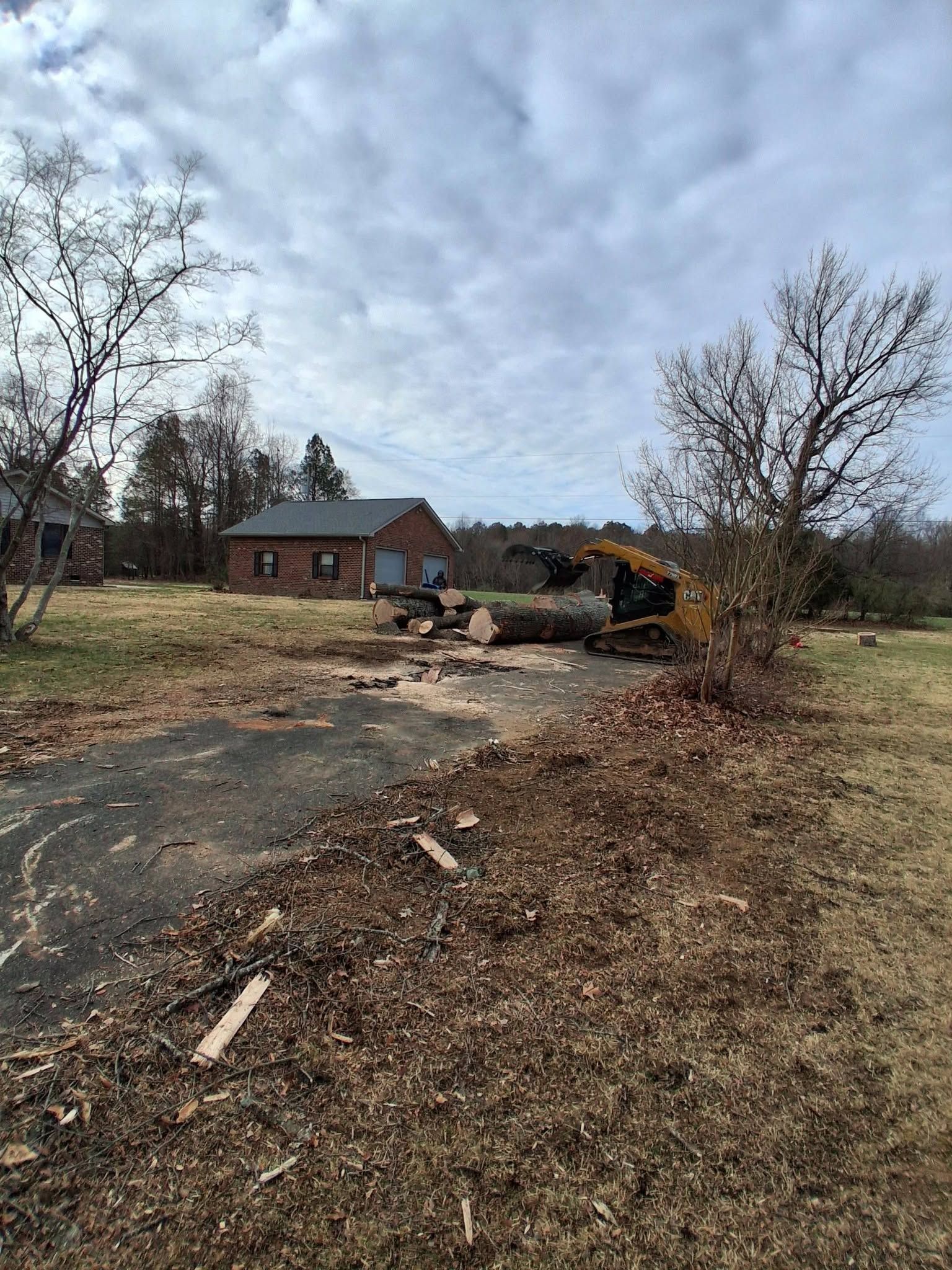 Rural driveway with debris and a damaged vehicle near a brick house under a cloudy sky