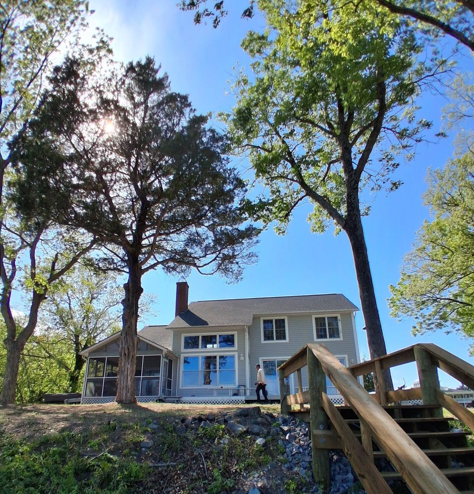 Two-story house on a wooded hill, seen from below with stairs in the foreground and blue sky above