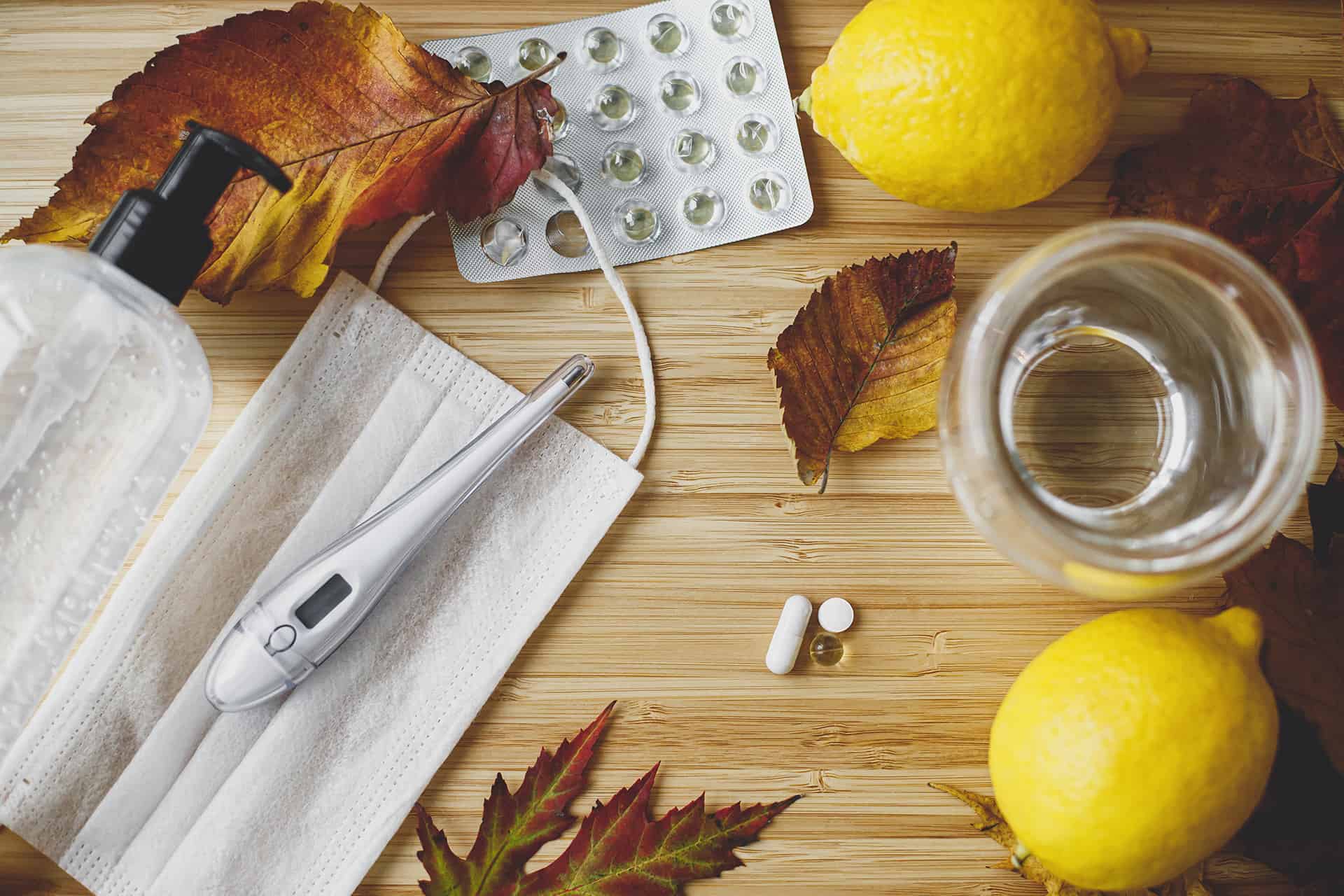 A tabletop collage of lemons, medication, blister packs, fall leaves, thermometers, masks, and hand sanitizer.
