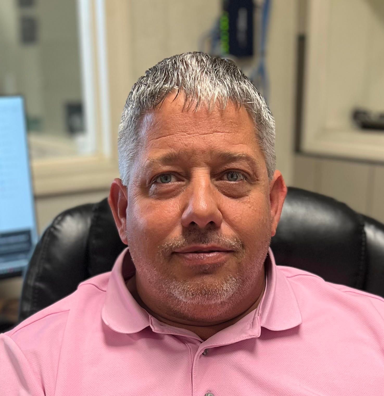 A man in a pink shirt is sitting in a chair in front of a computer.