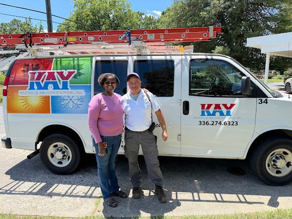 Two people standing beside a white KAY VAN work van with a ladder on top outdoors