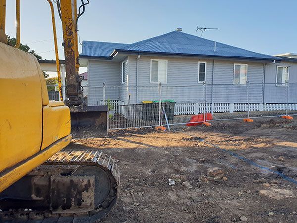 A yellow excavator is demolishing a house behind a fence.