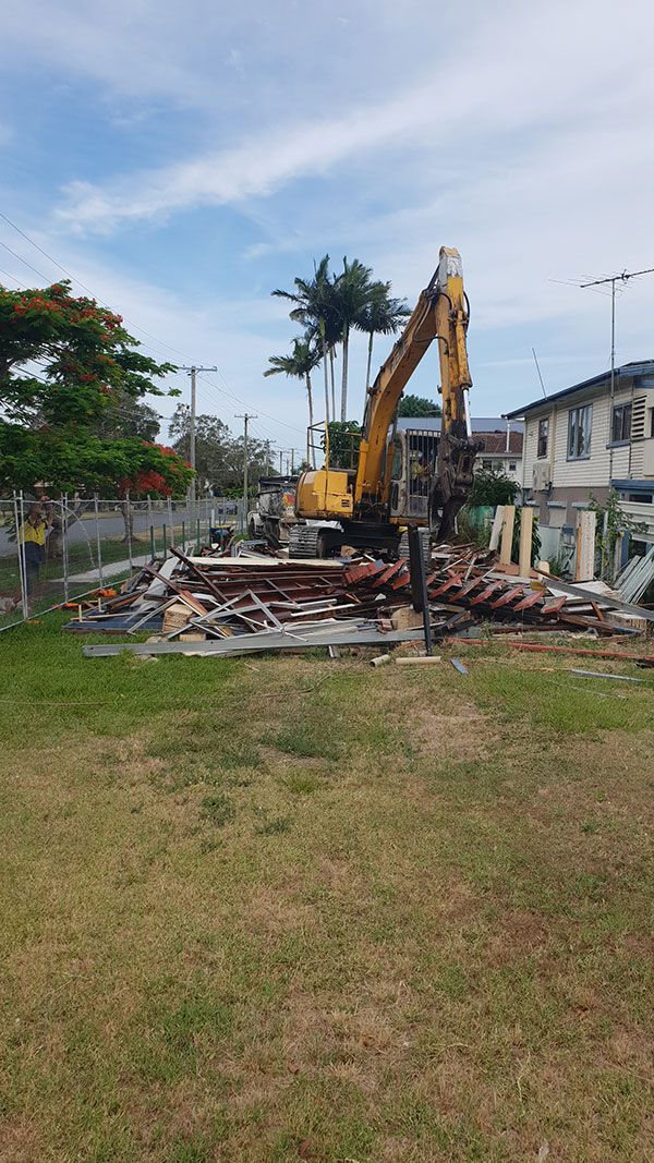 A yellow excavator is demolishing a house behind a fence.