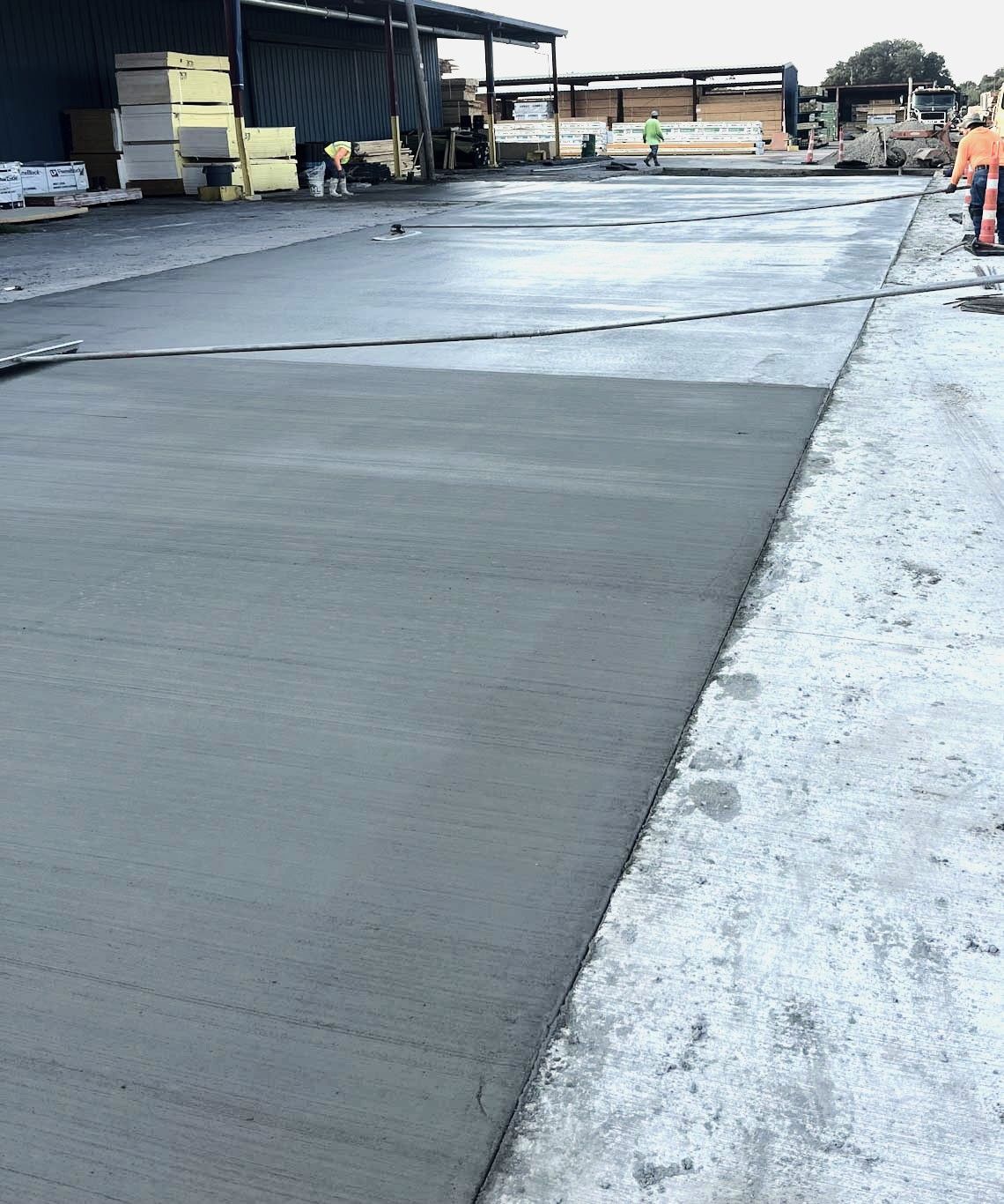 Freshly poured concrete surface, part of a larger construction site, with a worker in the background. Grey tones dominate.