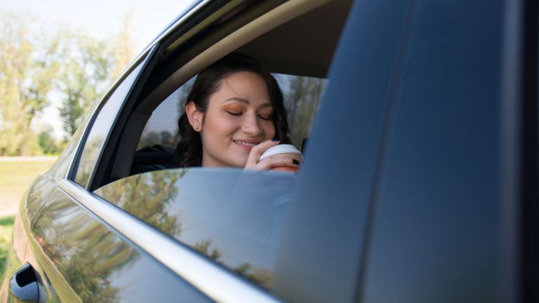 A woman is sitting in a car drinking coffee and talking on a cell phone.