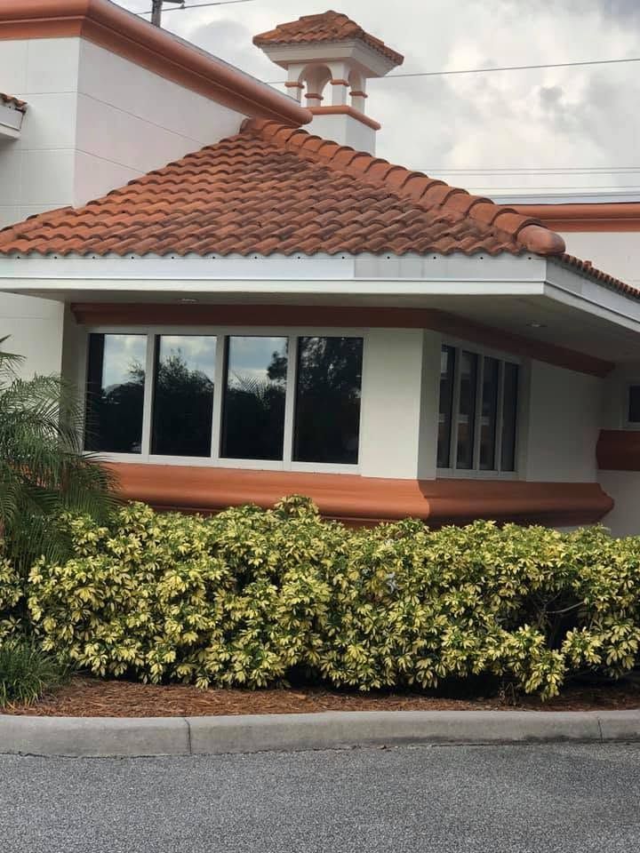 Stucco building with red tile roof, large windows, and trimmed hedge beside a driveway