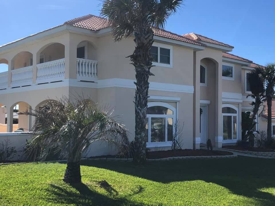 Large two-story beige house with red tile roof, white balconies, palm trees, and green lawn under a blue sky