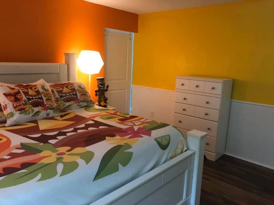 Bright bedroom with white bed, colorful floral bedding, lamp, and dresser against orange-yellow walls