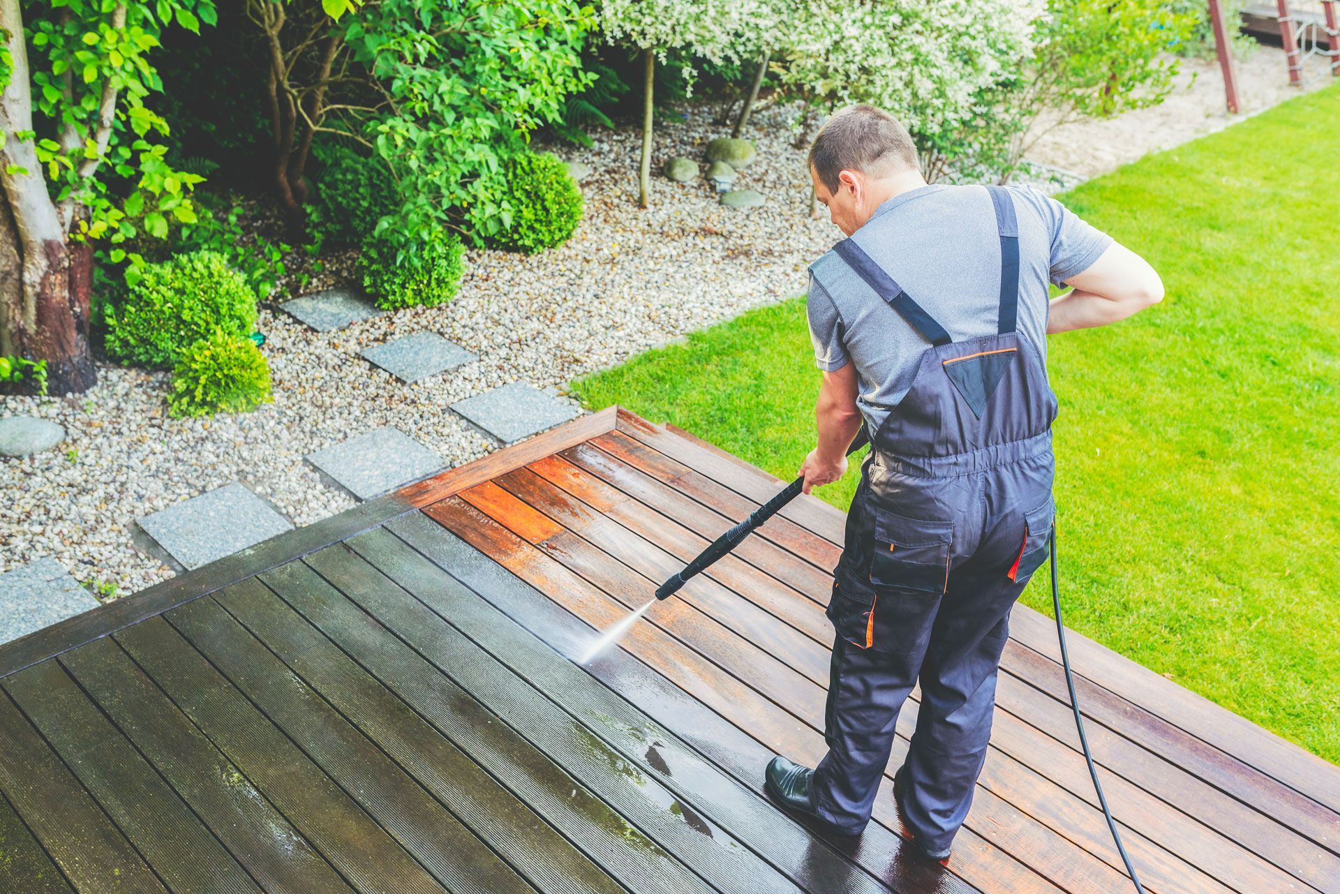 Person pressure-washing a wooden deck beside a green lawn and garden.