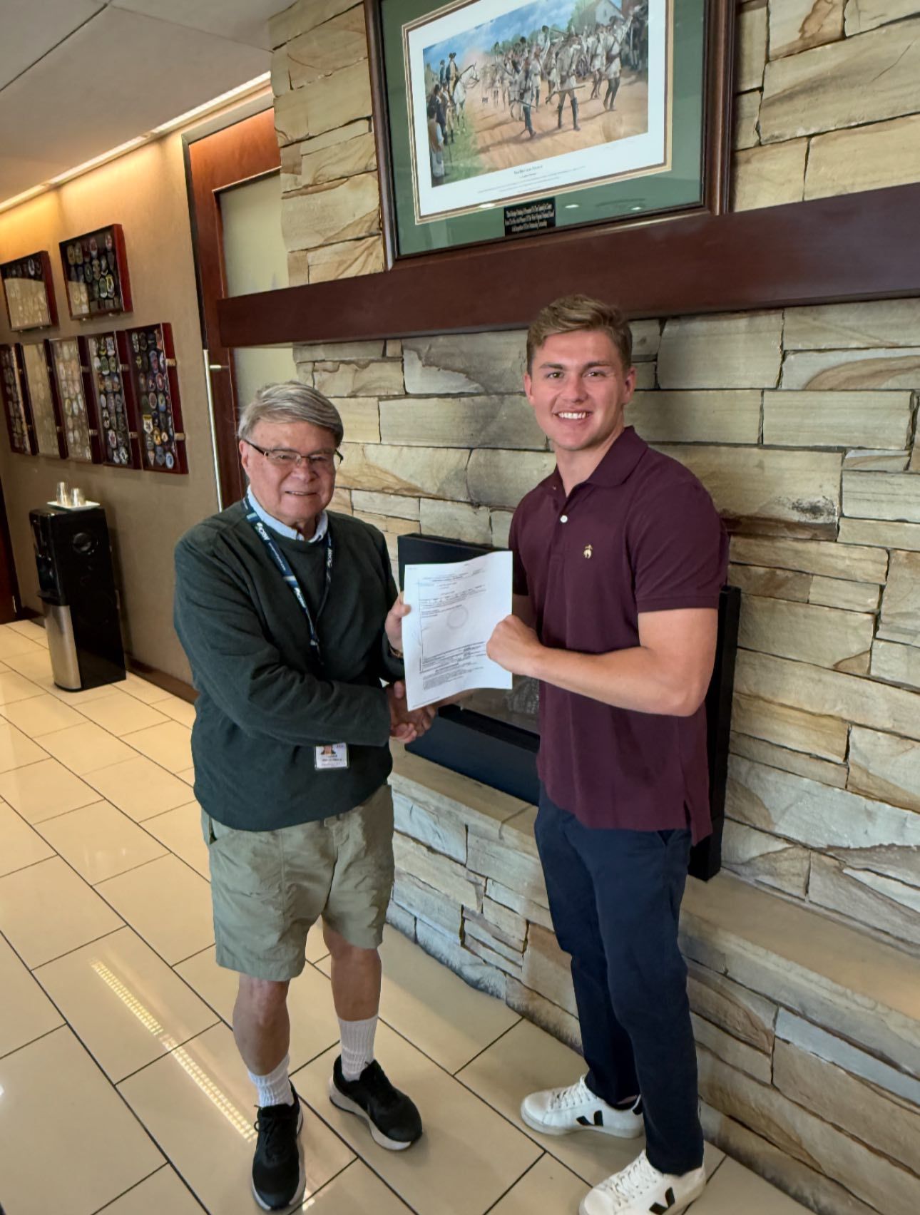 Two men shaking hands, one older in shorts, the other younger in a burgundy shirt; in a building by a fireplace.