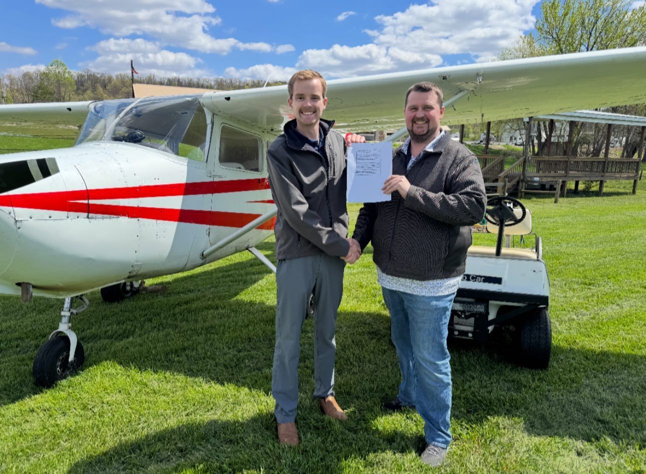 Two men shaking hands by a small airplane, one holding a paper. Outdoors, sunny day.