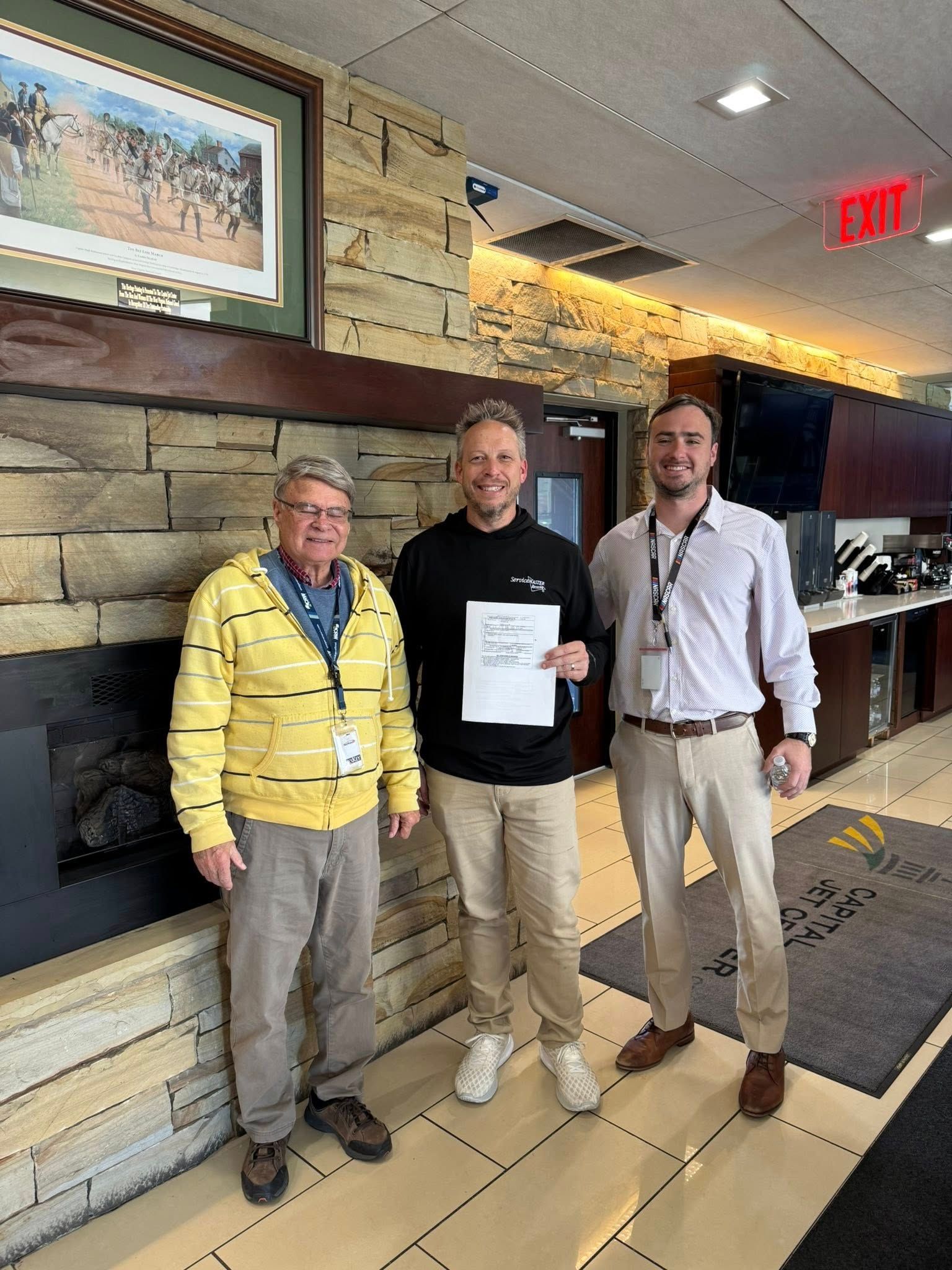 Three men standing inside, one holding a paper. The setting appears to be a lobby with a stone fireplace and art on the wall.