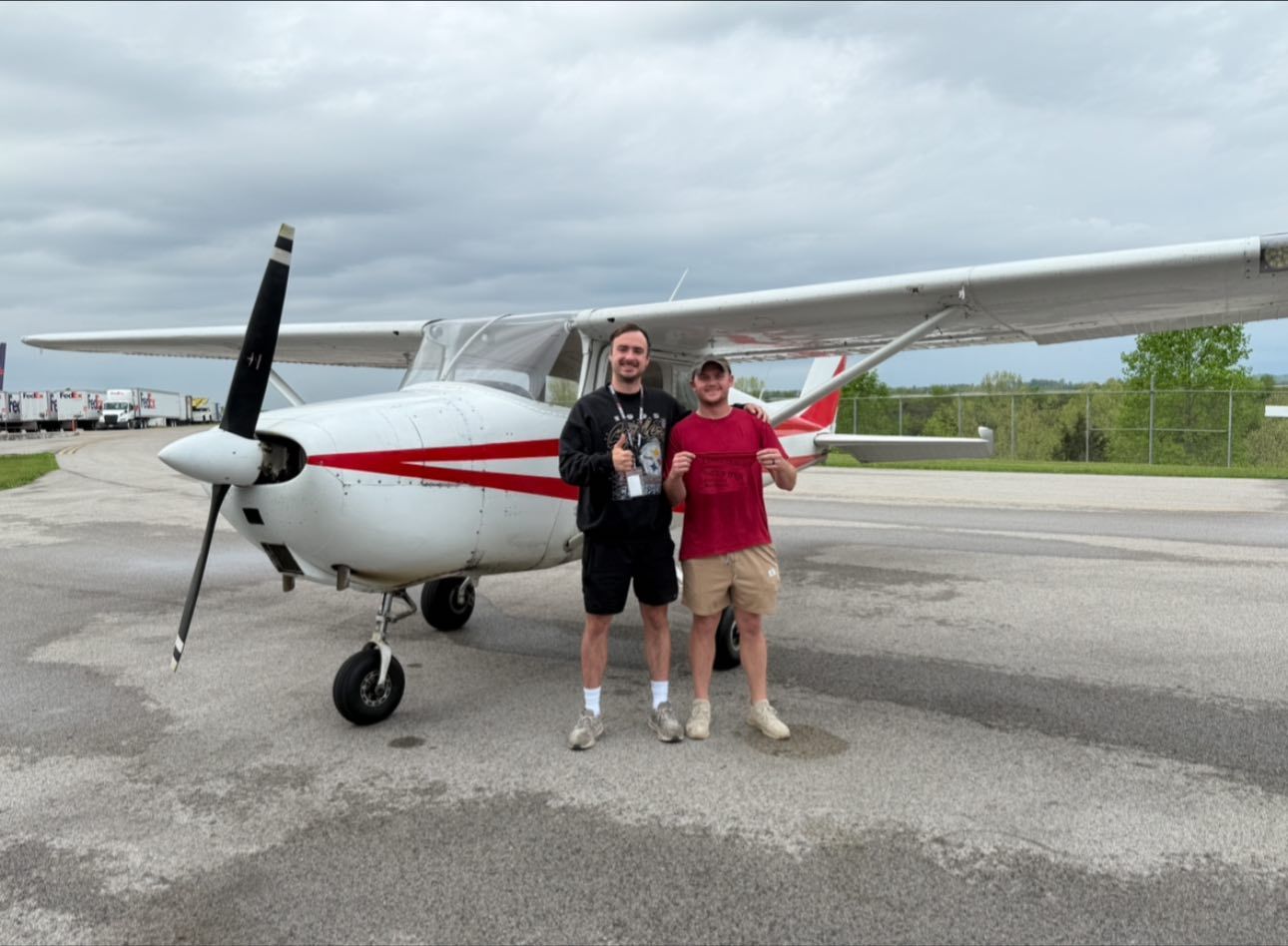 Two men stand next to a white and red airplane on a tarmac. One gives a thumbs up. Cloudy sky.