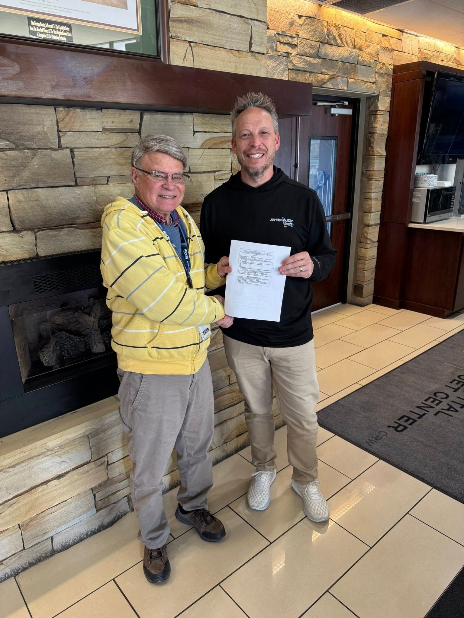 Two men smiling, shaking hands indoors, holding a paper, by a fireplace.