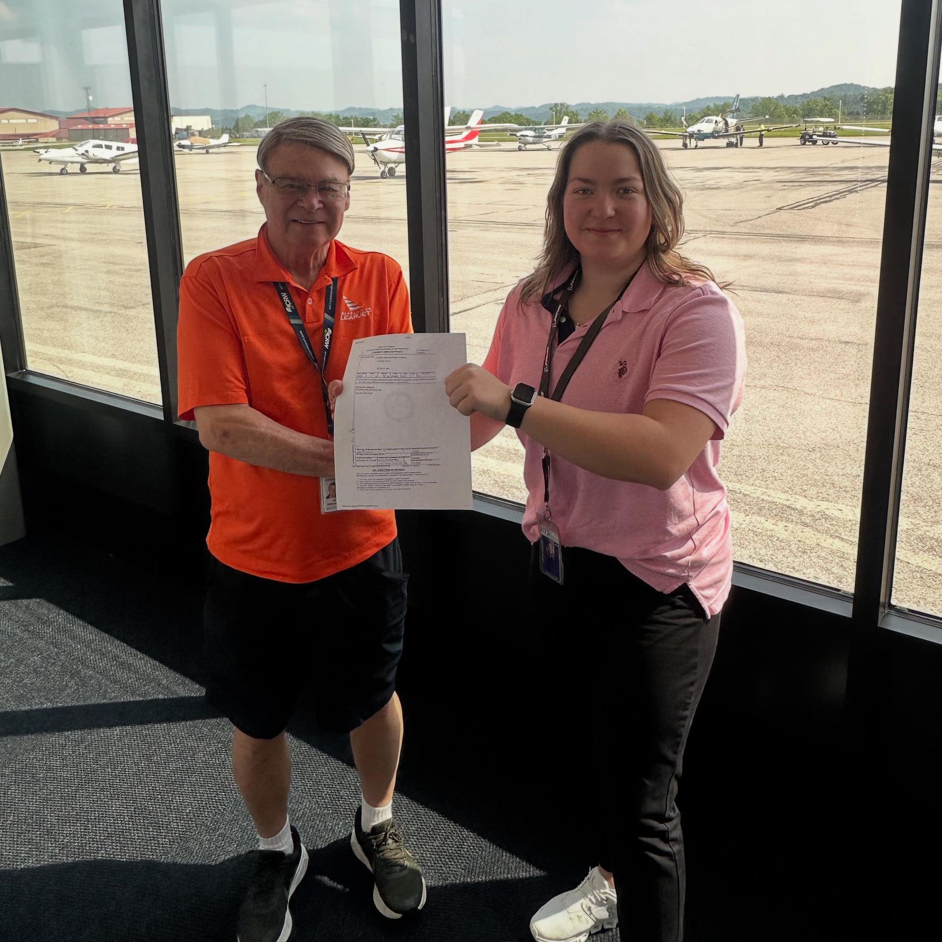 Man in orange shirt hands document to woman in pink shirt at airport. Airport view in background.