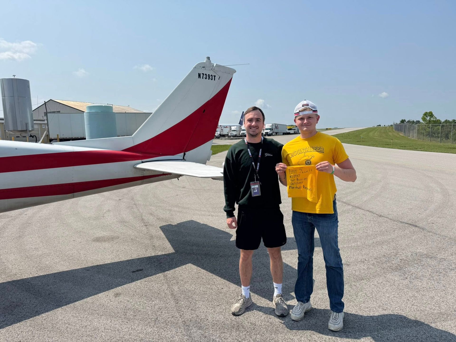 Two men stand near a small plane on a tarmac. One holds a yellow bag. Sunny day.
