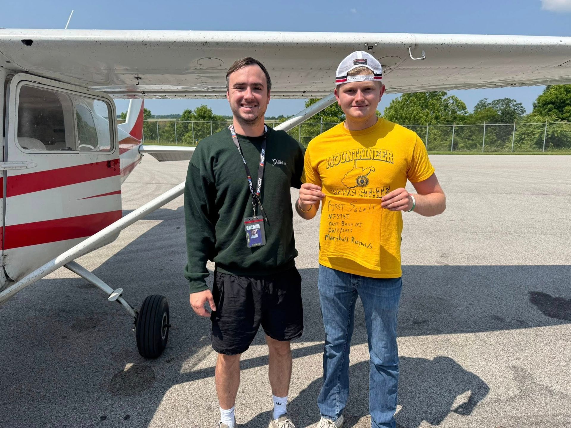Two men stand by a small airplane. One wears a green sweatshirt, the other a yellow shirt and jeans. They hold a piece of paper.