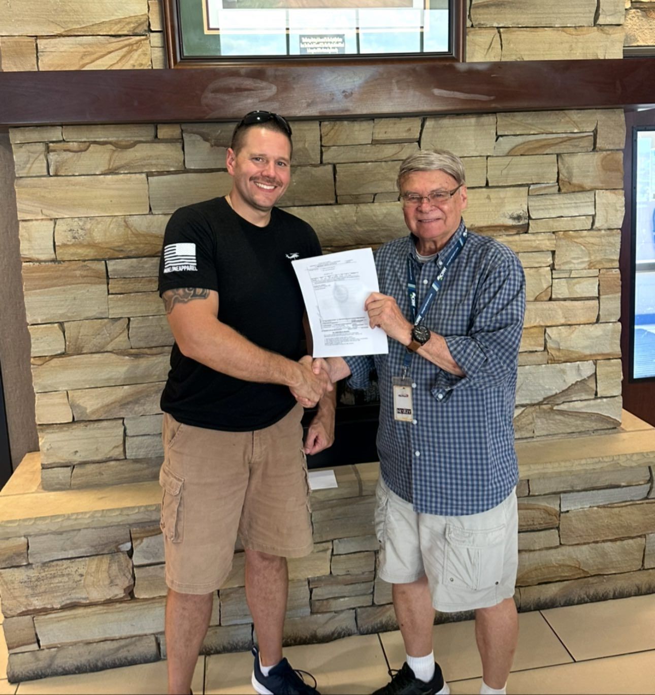 Two men shaking hands; a signing. One wears a black t-shirt and khaki shorts. The other holds a paper and is wearing a plaid shirt and shorts.