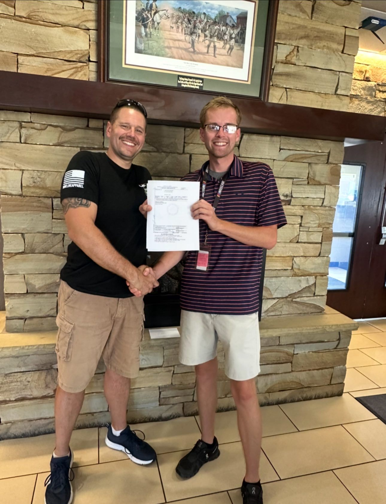 Two men shaking hands, one holding a document. Standing in a brick and wooden interior.