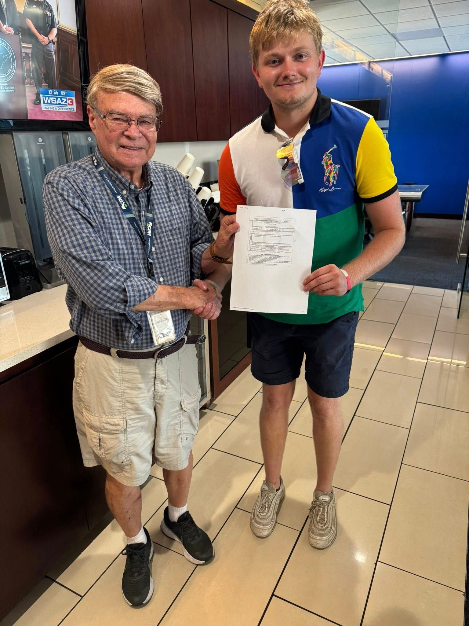 Two men shaking hands. One in shorts, a colorful polo shirt, and the other in a button-down shirt and khaki shorts. Indoor setting.