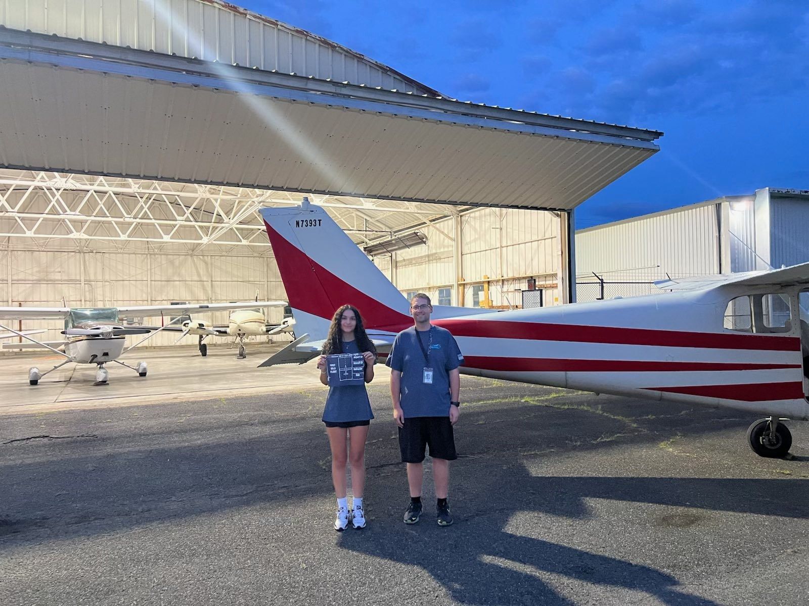 Teen and man stand by a small plane at dusk, hangar in background.