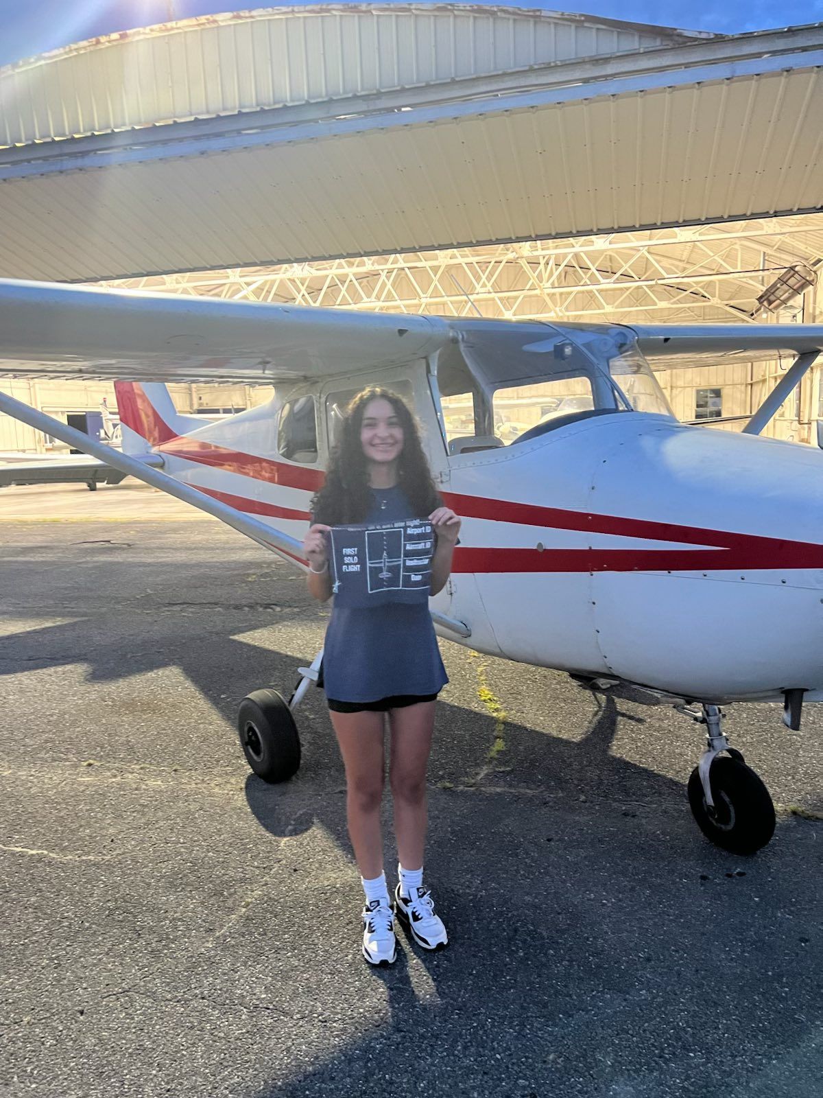 Young woman stands in front of a small airplane on a sunny tarmac, holding a sign.