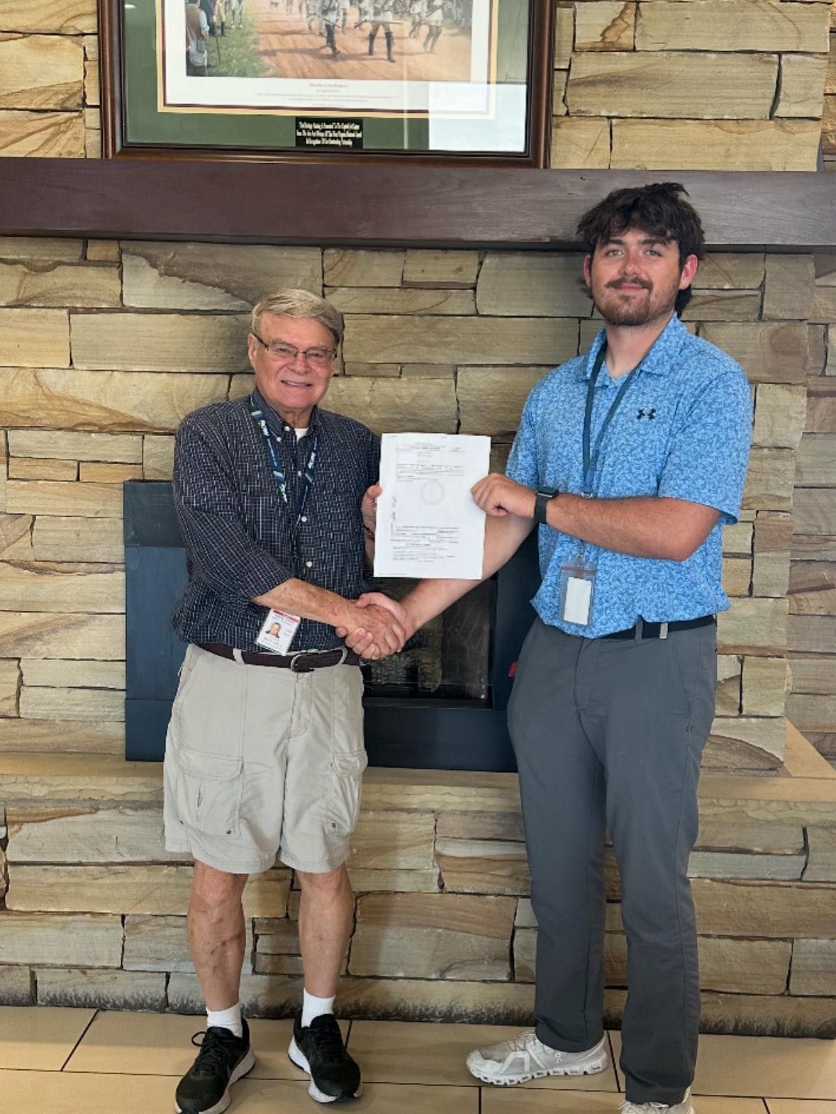 Two men shaking hands near a fireplace. One holds a paper, smiling. Background is a stone wall.