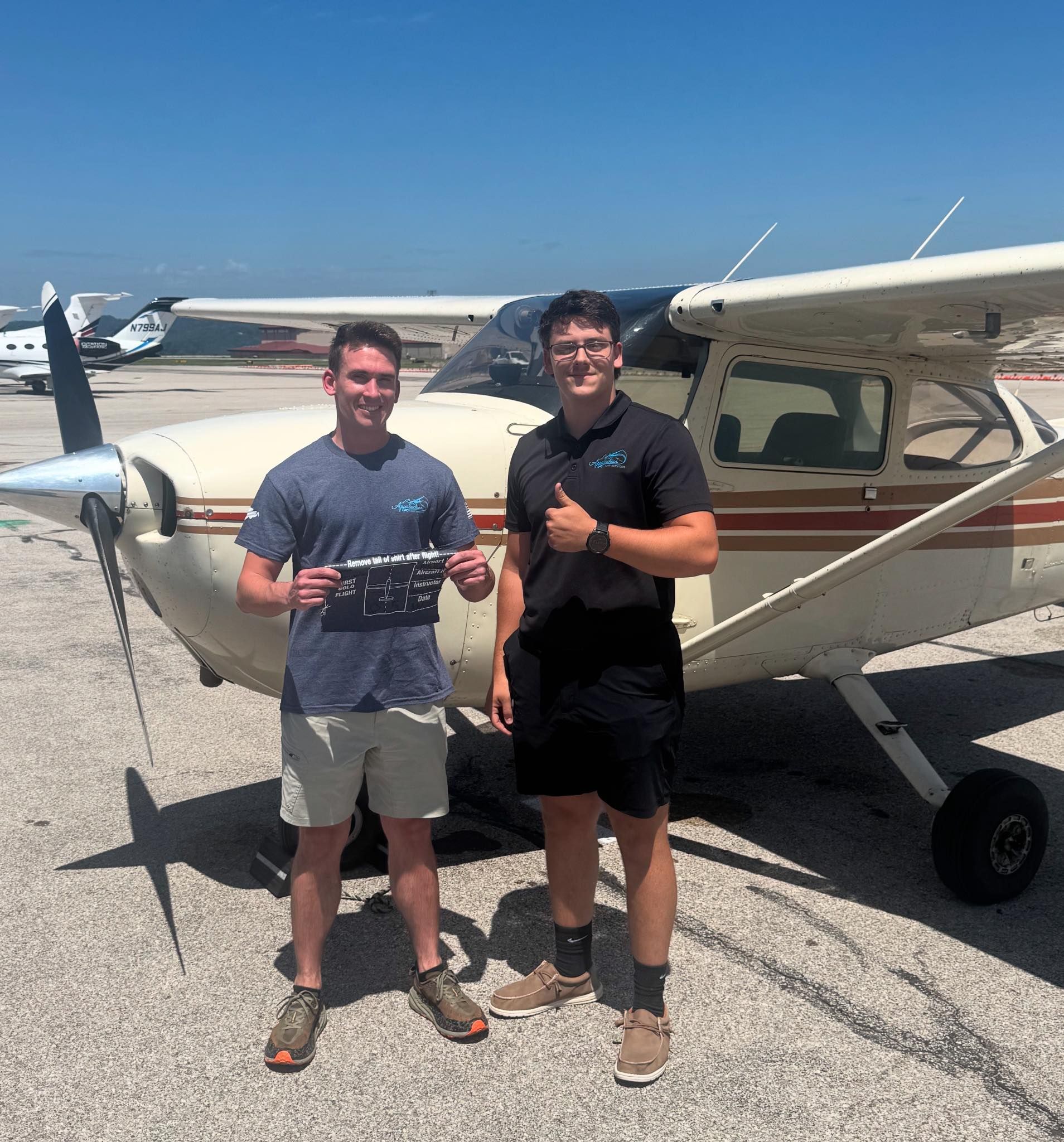 Two men stand by a small plane on a sunny airfield, one holding a plaque, the other giving a thumbs up.