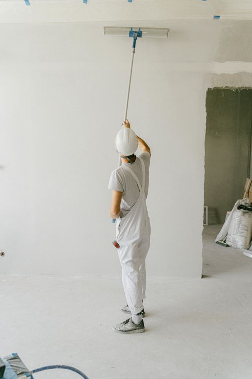 Person in white overalls and hard hat using a pole tool on a white ceiling during construction.