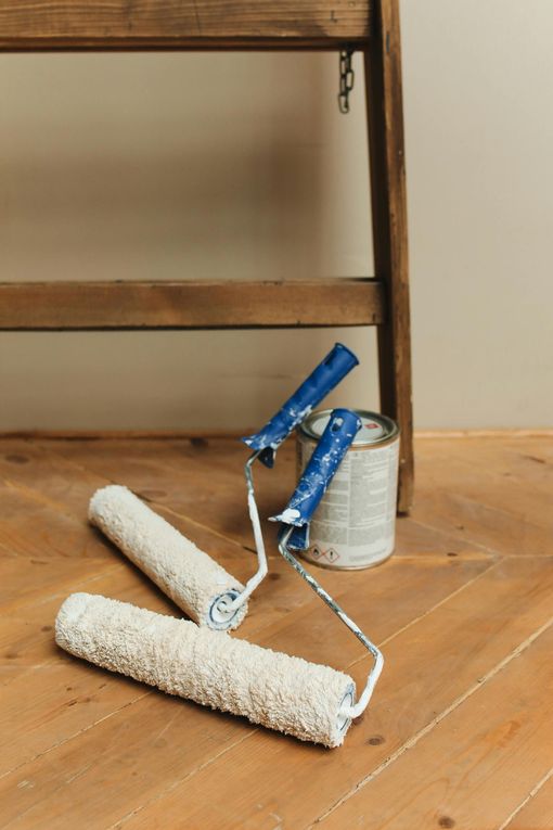 Paint rollers and a paint can on a wooden floor, near a brown ladder.
