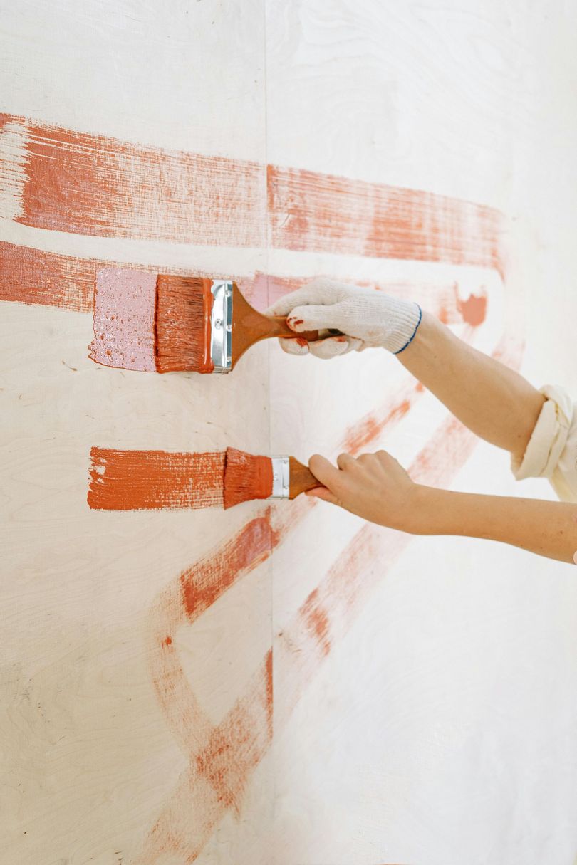 Two hands painting a white wall with a rusty orange-red paint using paintbrushes.