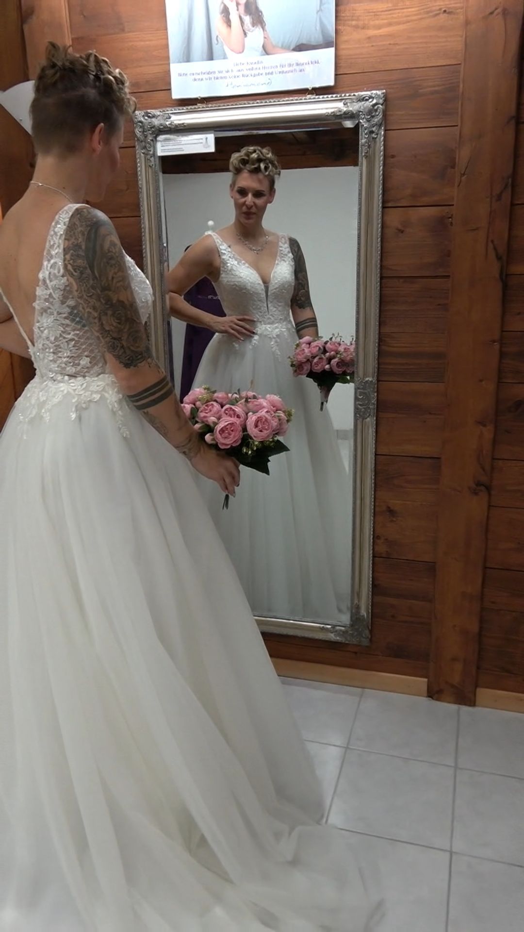 Eine Frau in einem Brautkleid steht vor einem Spiegel und hält einen Blumenstrauß in der Hand.