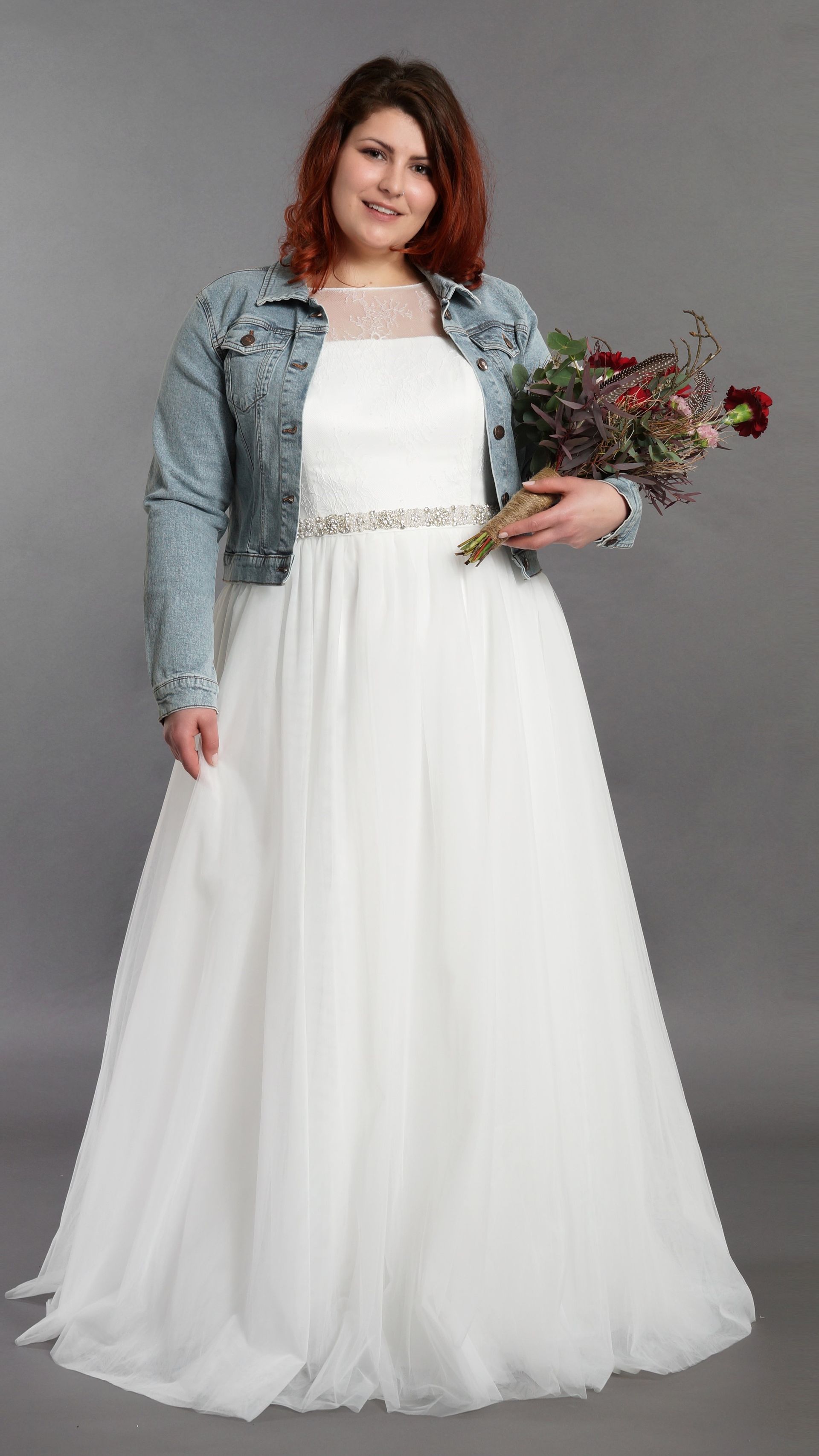 Eine Frau in einem weißen Brautkleid und einer Jeansjacke hält einen Blumenstrauß in der Hand.
