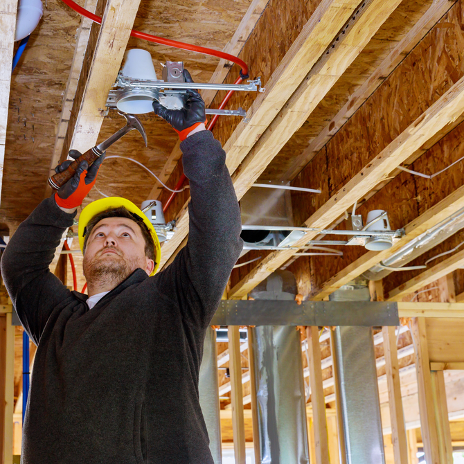 A construction worker in a hard hat hammers a ceiling light fixture in a building's unfinished frame.