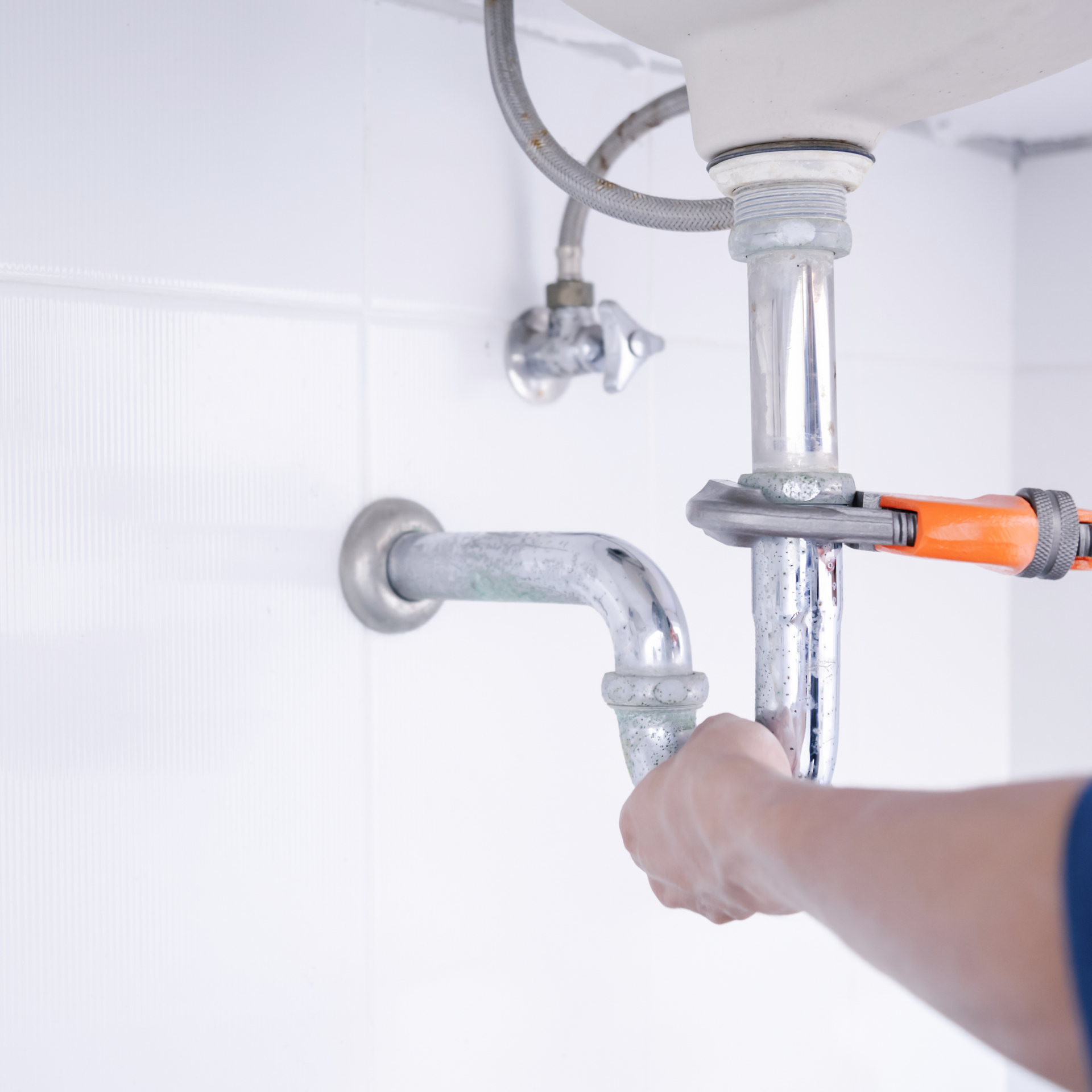Plumber using a wrench on a sink pipe in a white tiled bathroom.