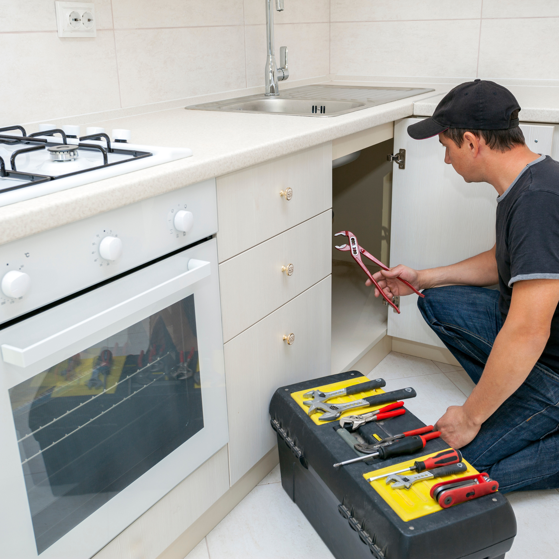 Man kneeling, working on plumbing under kitchen sink, toolbox open nearby.