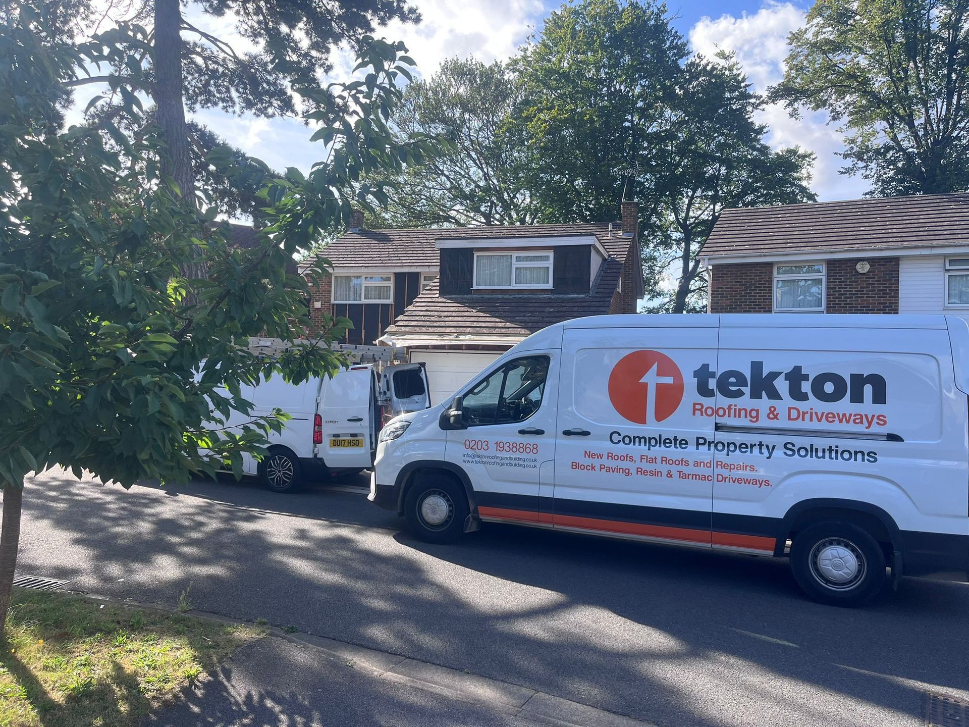 Two white Tekton roofing ang building vans parked on a residential street in front of houses with trees and blue sky.
