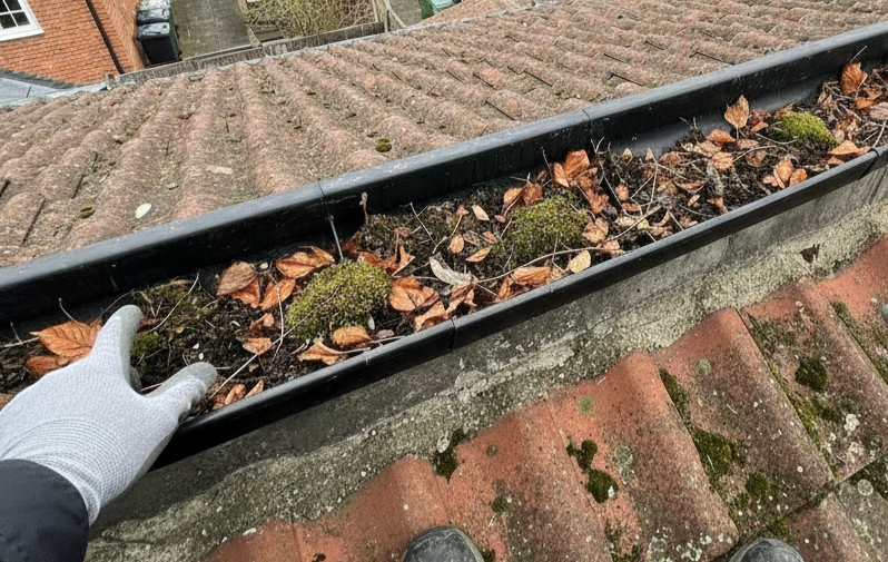 Gloved hand pointing to a debris-filled gutter on a tiled roof in Isleworth, with moss and leaves visible.