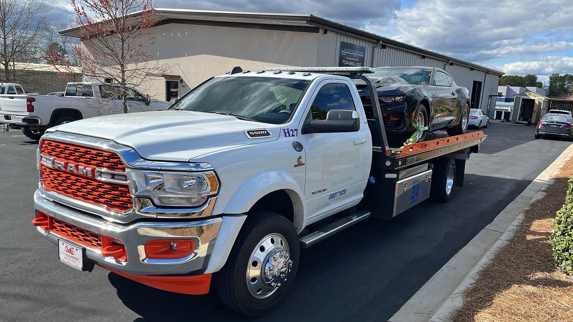 White tow truck with orange accents carrying a black car. The truck is parked outside a building on a sunny day.