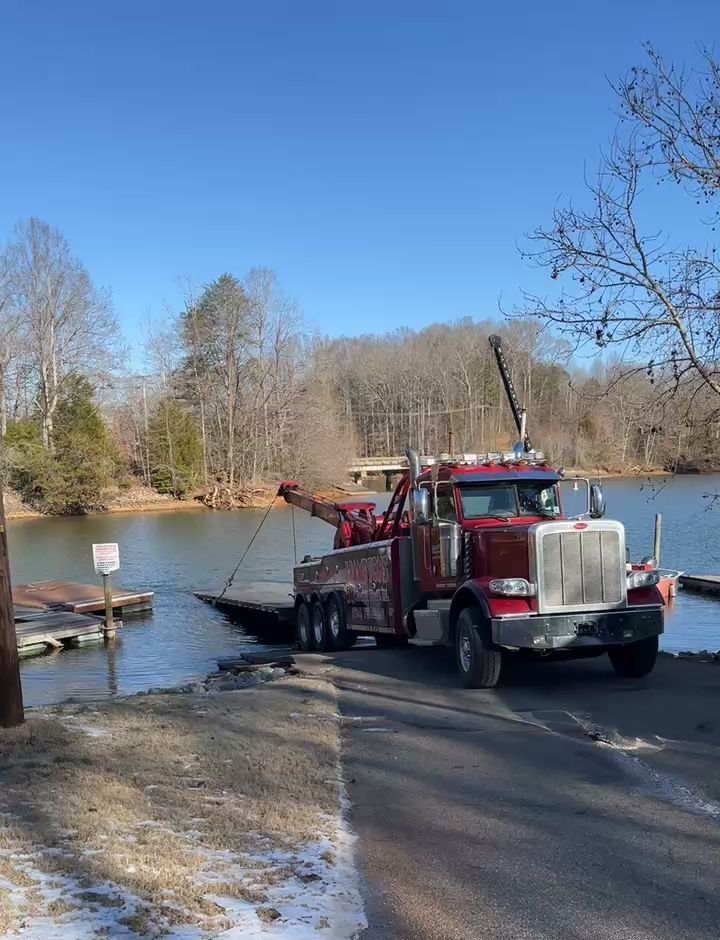 A red tow truck pulls a dock out of a body of water. Trees line the distant shore under a blue sky.
