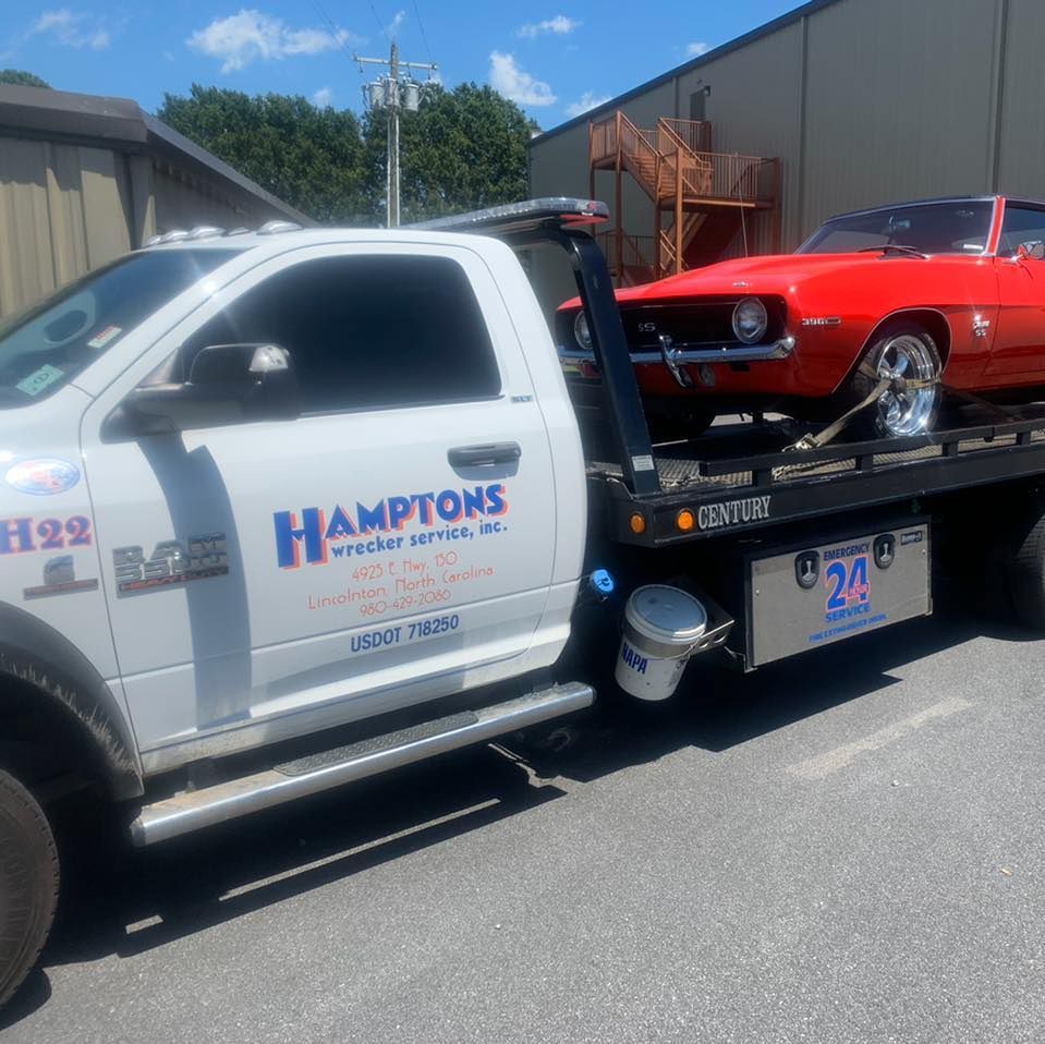 A red classic car is towed on a flatbed truck by Hamptons Wrecker Service on a sunny day.