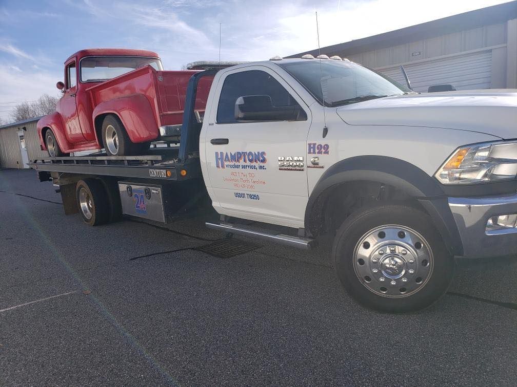 A red classic pickup truck being transported on the flatbed of a white tow truck in a paved lot.