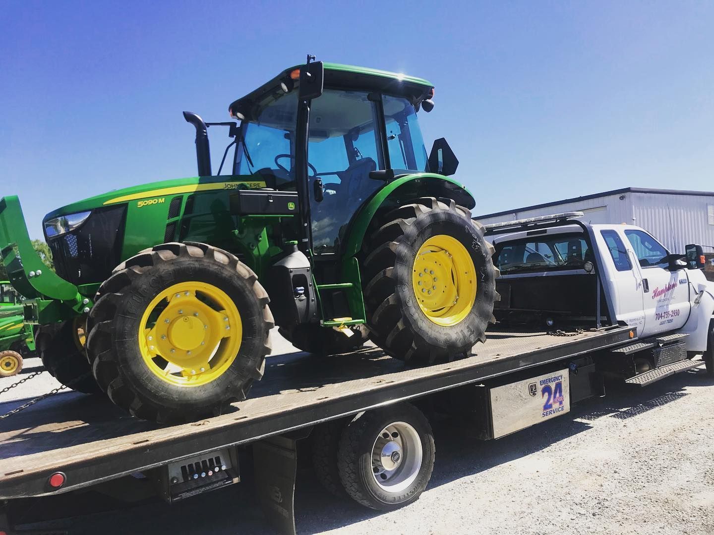 A green and yellow John Deere tractor loaded on a white flatbed tow truck under a clear blue sky.