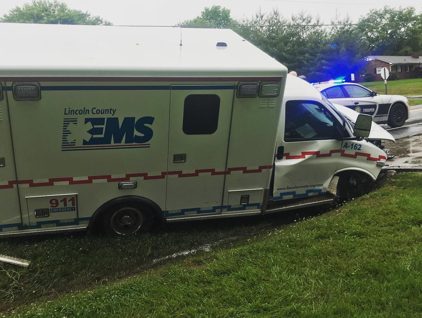 An ambulance stuck in a grassy ditch with a damaged front. A police car with flashing blue lights is in the background.