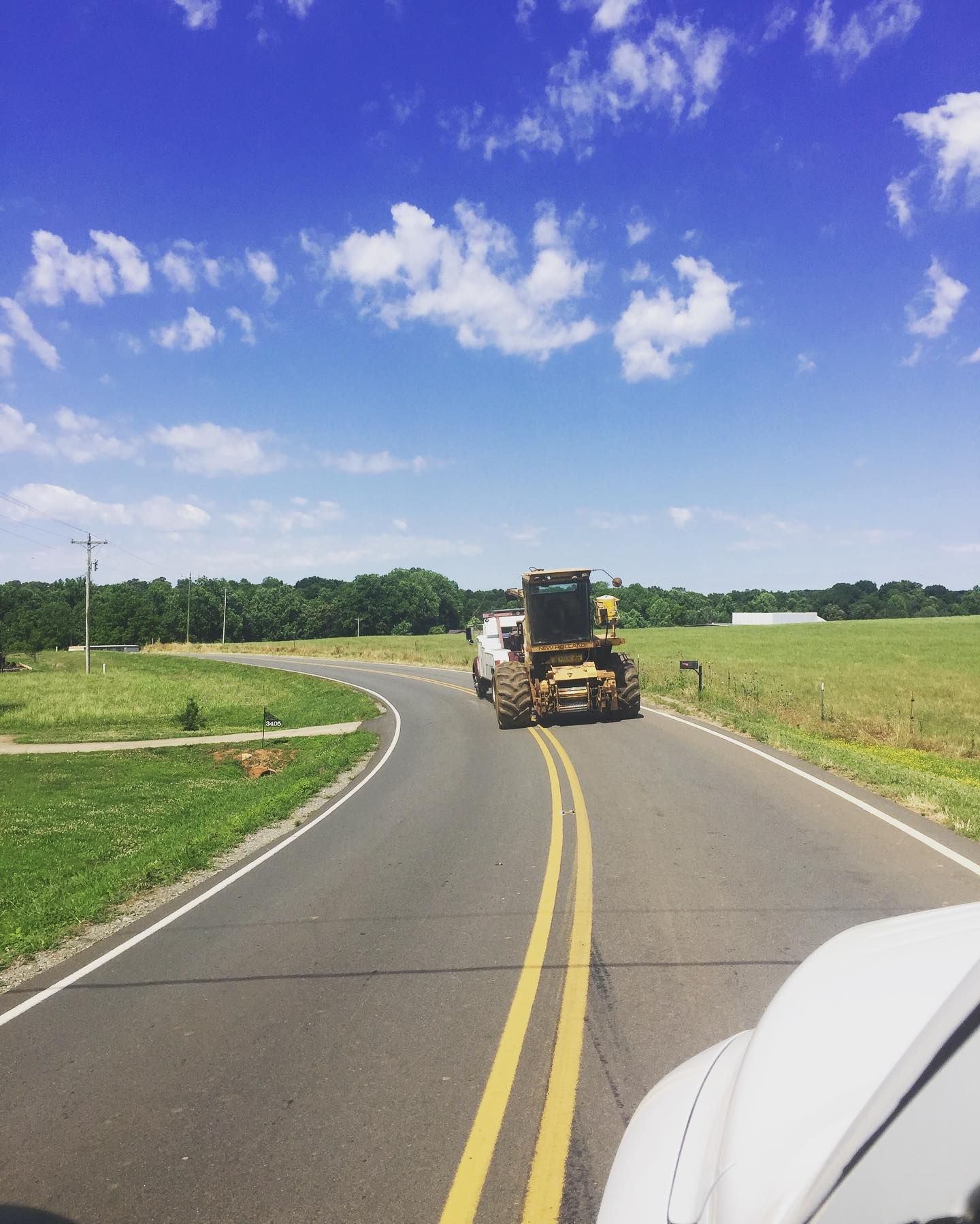 A truck towing construction equipment on a curved road in a rural setting under a blue sky with clouds.