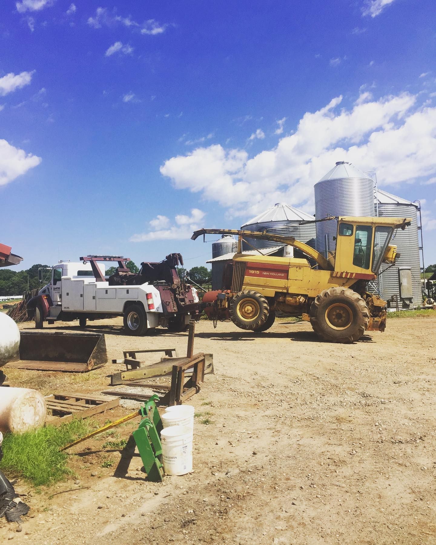 A white tow truck is attached to a yellow farm machine in a rural setting with silos under a bright blue sky.