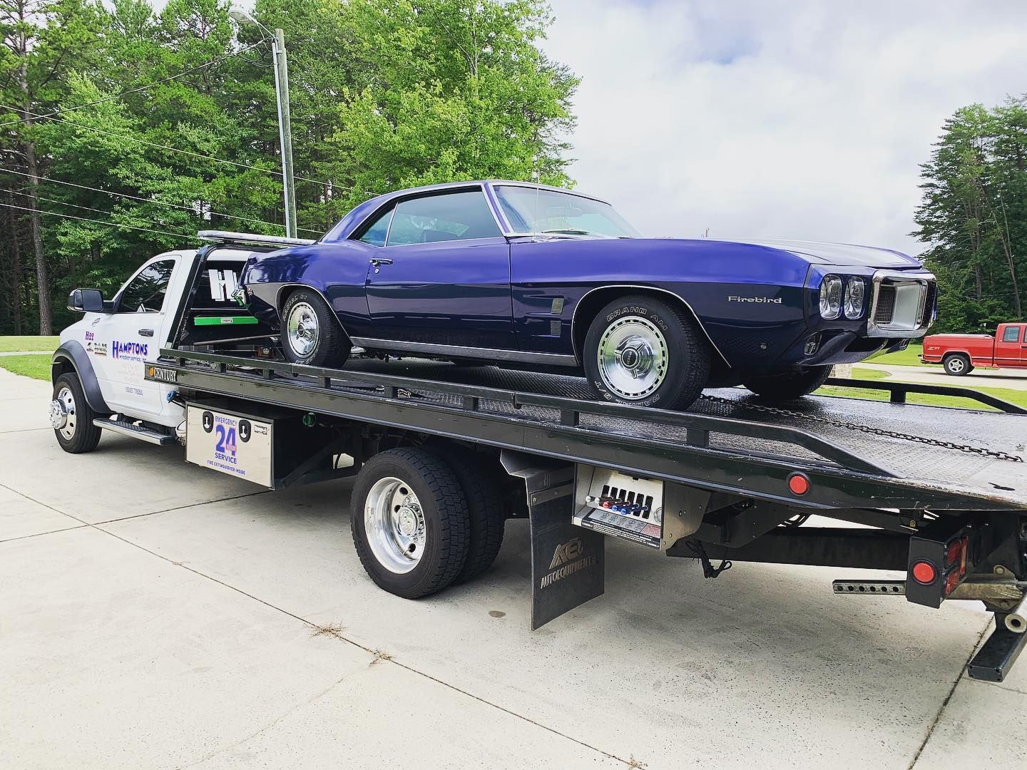 A classic blue Pontiac Firebird is being transported on a flatbed tow truck, outdoors on a sunny day.